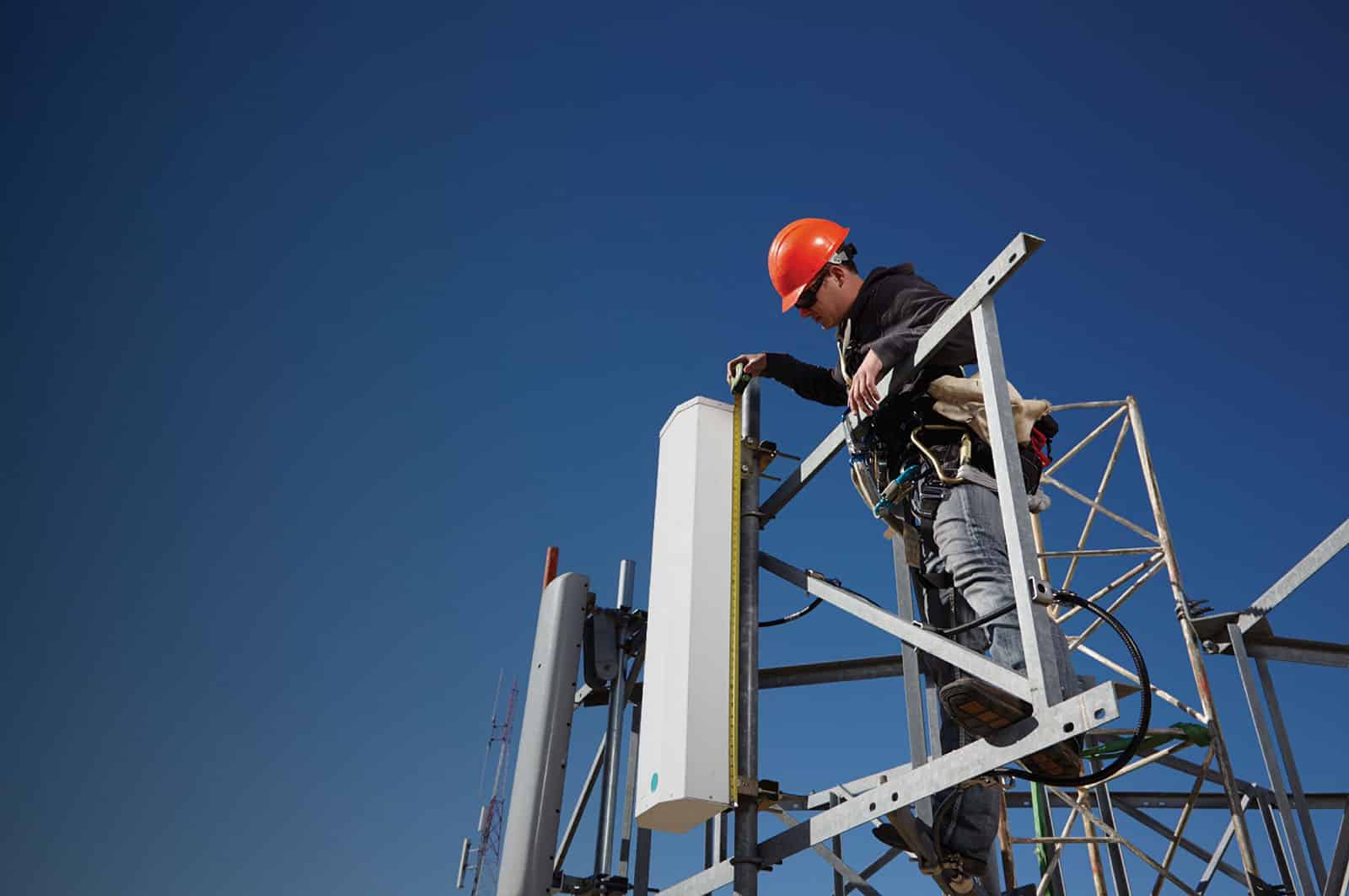 antenna A construction worker wearing an orange hard hat and safety gear stands on a metal scaffolding, working on a high structure against a clear blue sky.