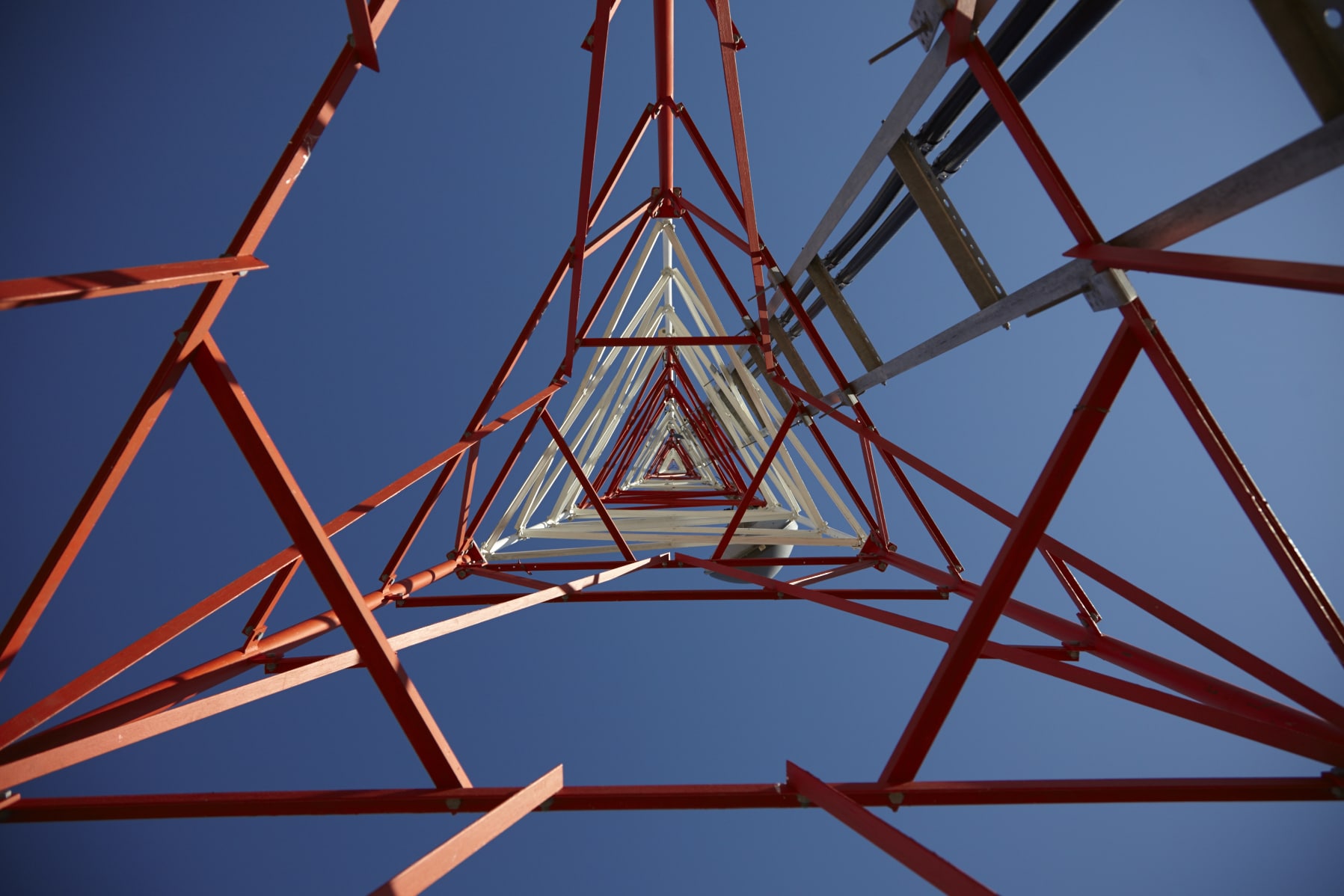 bt-758 View from beneath a tall red and white metal transmission tower, looking up into a clear blue sky. The geometric patterns formed by the structure's crossbeams create a series of symmetrical triangular shapes.